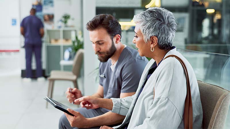 Profissional de saúde masculino com bata cinzento e cliente sénior de casaco branco sentados lado a lado numa sala de espera hospitalar, a analisar informação num tablet digital